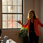 Woman in a pink blazer giving a presentation in a sunlit room, with a projection screen to her right and attendees in the foreground.