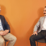 Two men in suits sit facing each other in discussion against an orange background.