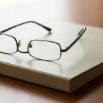Metal-framed eyeglasses rest on a closed white notebook on a wooden desk.