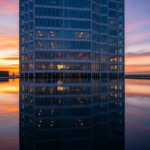 Skyscraper with glass façade at sunset, its lit windows reflected in a calm rooftop pool by the waterline.