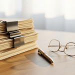 Pile of manila file folders clipped with metal binder clips on a desk, with glasses and a pen nearby