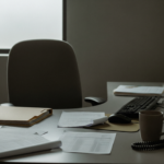 Office desk scene with papers, a mug, keyboard, mouse, monitor, and a phone; empty chair with a window in the background, dim lighting.