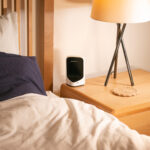 Bedside scene with a white lamp on a wooden nightstand and a small white device beside a navy bedspread and pillow contrast.