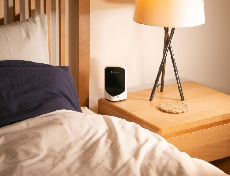 Bedside scene with a white lamp on a wooden nightstand and a small white device beside a navy bedspread and pillow contrast.