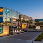 Modern glass office complex with a teal logo, palm trees, and fountain jets at dusk/dawn.