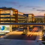 Modern office campus at dusk with glass buildings, warm interior lights, and a central entrance logo.