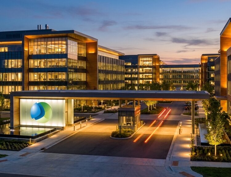 Modern office campus at dusk with glass buildings, warm interior lights, and a central entrance logo.