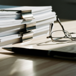 Stack of papers clipped with metal binder clips on a desk, with reading glasses nearby.
