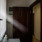 Clinic corridor corner with a looped hanging cord, a medical light on the left, and an open wooden door ahead.