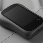 Compact black touchscreen device resting on smooth gray fabric, with a circular button on the right edge.