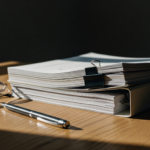 Stack of printed documents clipped with binder clips on a wooden desk, with glasses and a pen nearby.